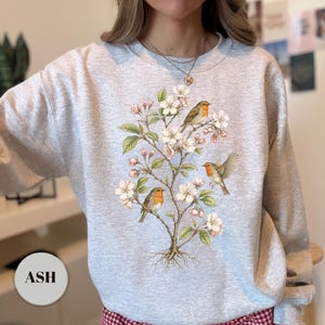 May include: Ash-colored sweatshirt featuring a detailed illustration of a flowering tree with three robins. The design includes white blossoms, green leaves, and the birds with orange breasts. The word "ASH" is printed on a circular label.