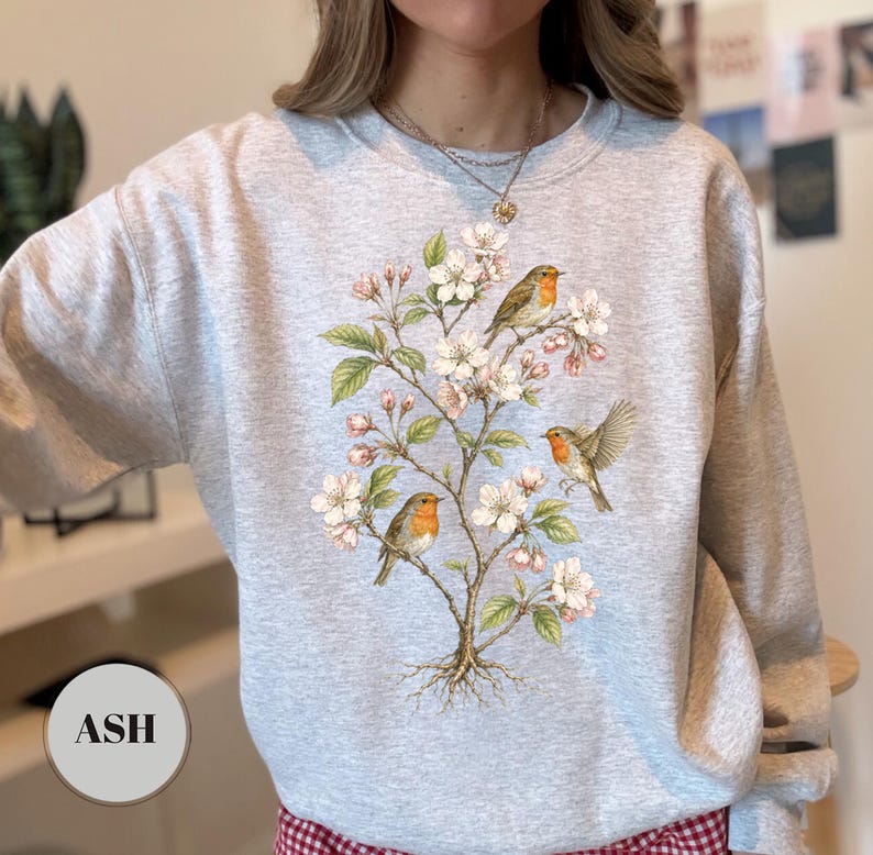 May include: Ash-colored sweatshirt featuring a detailed illustration of a flowering tree with three robins. The design includes white blossoms, green leaves, and the birds with orange breasts. The word "ASH" is printed on a circular label.