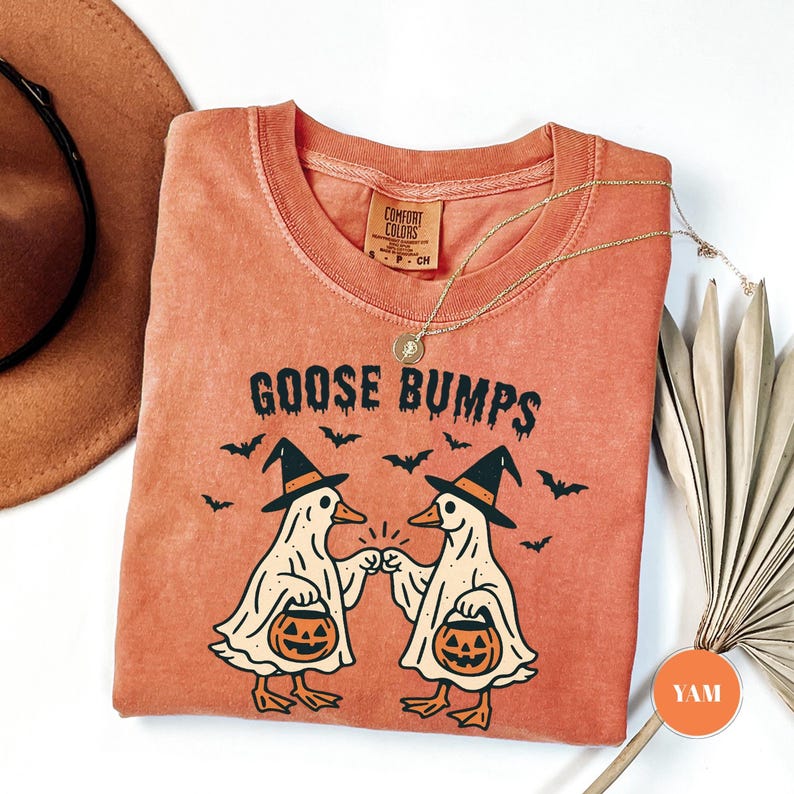 May include: An orange Comfort Colors t-shirt with the words "GOOSE BUMPS" and a graphic of two cartoon geese wearing witch hats and holding jack-o'-lanterns. Bats are flying above the geese. A gold necklace is draped across the shirt.