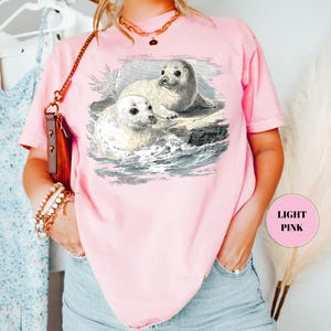 Pode incluir: T-shirt rosa claro com uma ilustra&ccedil;&atilde;o vintage de duas focas numa rocha, com ondas ao fundo. As focas s&atilde;o representadas em tons de cinza e branco. A t-shirt tem mangas curtas e um corte folgado. O texto "LIGHT PINK" &eacute; vis&iacute;vel.