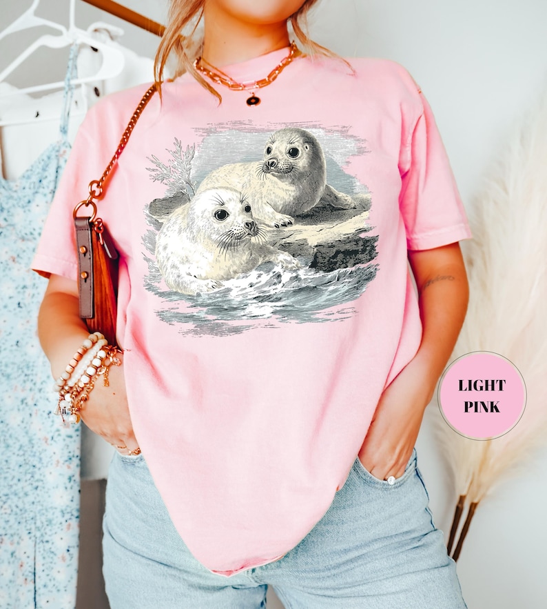Pode incluir: T-shirt rosa claro com uma ilustra&ccedil;&atilde;o vintage de duas focas numa rocha, com ondas ao fundo. As focas s&atilde;o representadas em tons de cinza e branco. A t-shirt tem mangas curtas e um corte folgado. O texto "LIGHT PINK" &eacute; vis&iacute;vel.