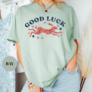 K&ouml;nnte beinhalten: Hellgr&uuml;nes T-Shirt mit dem Schriftzug "GOOD LUCK" in Marineblau &uuml;ber einer roten Hasengrafik. Das Design beinhaltet Hufeisen und Kleebl&auml;tter. Das Shirt wird mit einer blauen Jeans kombiniert.