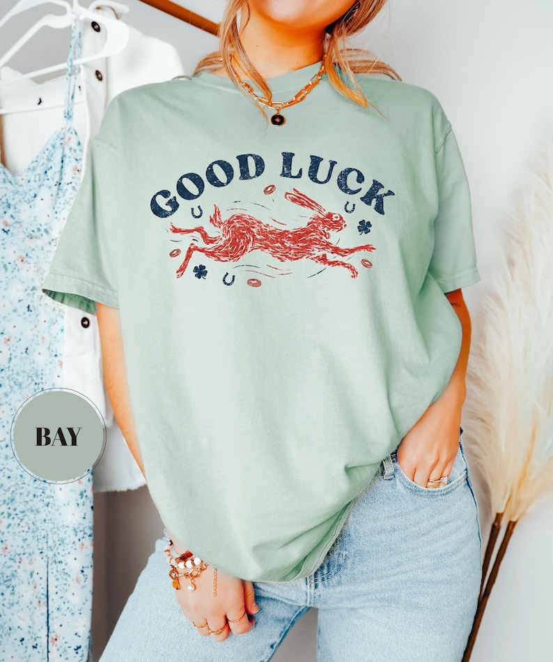 K&ouml;nnte beinhalten: Hellgr&uuml;nes T-Shirt mit dem Schriftzug "GOOD LUCK" in Marineblau &uuml;ber einer roten Hasengrafik. Das Design beinhaltet Hufeisen und Kleebl&auml;tter. Das Shirt wird mit einer blauen Jeans kombiniert.