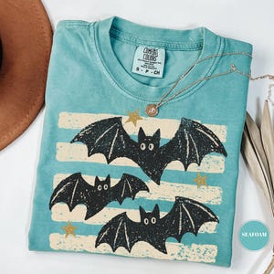 May include: A seafoam green short-sleeve t-shirt with a crew neckline and a Comfort Colors brand label. The graphic features three black bats flying over horizontal cream-colored distressed stripes, accented by small gold stars scattered throughout.
