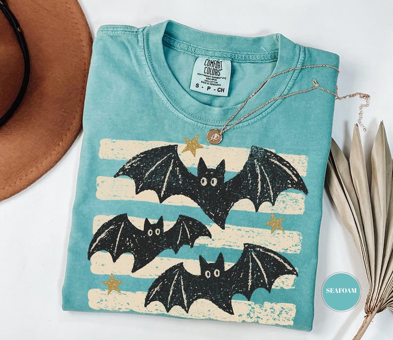 May include: A seafoam green short-sleeve t-shirt with a crew neckline and a Comfort Colors brand label. The graphic features three black bats flying over horizontal cream-colored distressed stripes, accented by small gold stars scattered throughout.