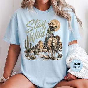 May include: Pale blue t-shirt with a desert-themed graphic. The design features a cowgirl on horseback, a setting sun, and the words "Stay Wild". A cactus and desert landscape are also visible.