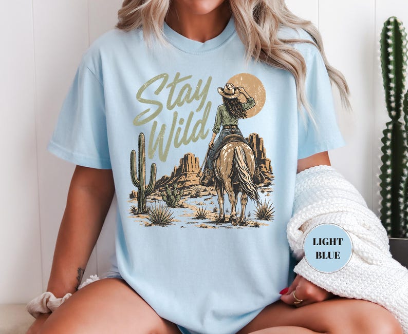 May include: Pale blue t-shirt with a desert-themed graphic. The design features a cowgirl on horseback, a setting sun, and the words "Stay Wild". A cactus and desert landscape are also visible.