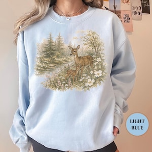 May include: Light blue sweatshirt featuring a detailed illustration of a deer and fawn in a forest setting. The artwork includes a river, trees, and white flowers. The sweatshirt has a crew neck and long sleeves. The text "LIGHT BLUE" is in a circle on the bottom right.