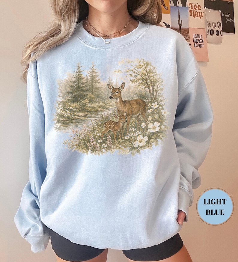 May include: Light blue sweatshirt featuring a detailed illustration of a deer and fawn in a forest setting. The artwork includes a river, trees, and white flowers. The sweatshirt has a crew neck and long sleeves. The text "LIGHT BLUE" is in a circle on the bottom right.