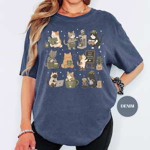 May include: Denim blue t-shirt featuring a whimsical illustration of cats engaged in various coffee-related activities. The cats are depicted reading, brewing coffee, and using laptops. The text "Coffee Makes Everything Possible" is on a small chalkboard.