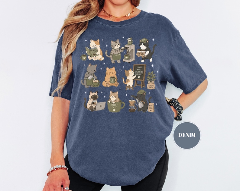 May include: Denim blue t-shirt featuring a whimsical illustration of cats engaged in various coffee-related activities. The cats are depicted reading, brewing coffee, and using laptops. The text "Coffee Makes Everything Possible" is on a small chalkboard.
