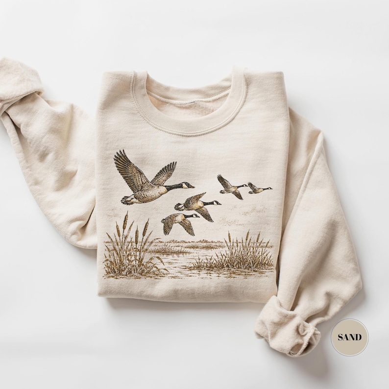 Peut inclure: Un sweat-shirt &agrave; col rond couleur sable orn&eacute; d'une illustration d&eacute;taill&eacute;e &agrave; l'encre brune repr&eacute;sentant un vol d'oies au-dessus d'un paysage mar&eacute;cageux avec des roseaux. Le motif est centr&eacute; et une &eacute;tiquette circulaire marqu&eacute;e SAND est visible.