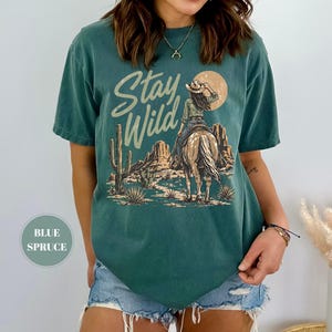 May include: Teal t-shirt with a desert-themed graphic. The design features a cowgirl on horseback with the text "Stay Wild" in a script font. A cactus and sun are also included in the design. The shirt has a relaxed fit.