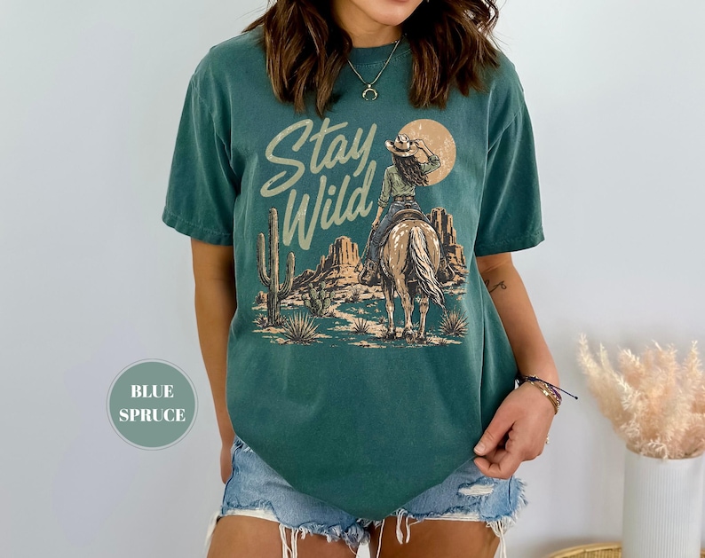 May include: Teal t-shirt with a desert-themed graphic. The design features a cowgirl on horseback with the text "Stay Wild" in a script font. A cactus and sun are also included in the design. The shirt has a relaxed fit.