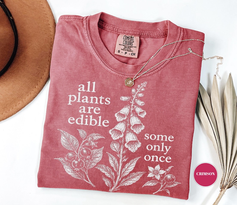 以下が含まれることがあります： くすんだピンク色の半袖Tシャツ。植物のイラストと「all plants are edible some only once」という文字が白でプリントされています。クルーネックで、襟の内側にはComfort Colorsのブランドラベルが付いています。