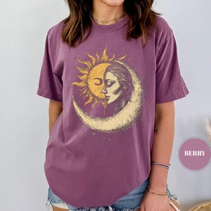 May include: Berry-colored t-shirt featuring a sun and moon graphic. The design depicts a sun with a face embracing a woman's face within a crescent moon. The shirt has short sleeves and a relaxed fit, perfect for casual wear.