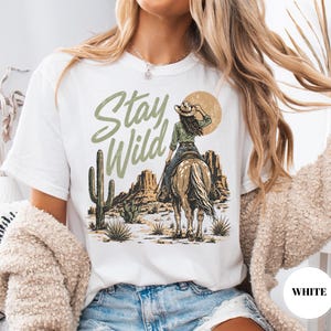 May include: White t-shirt with a desert-themed graphic. The design features a cowgirl on horseback, cacti, and the words "Stay Wild" in a green, distressed font. The shirt has a classic crew neck.