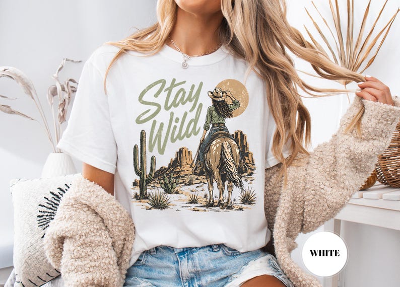 May include: White t-shirt with a desert-themed graphic. The design features a cowgirl on horseback, cacti, and the words "Stay Wild" in a green, distressed font. The shirt has a classic crew neck.