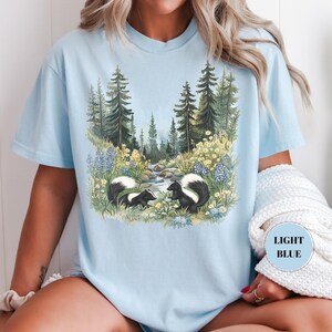 May include: Light blue t-shirt featuring a nature scene with two skunks near a stream, surrounded by wildflowers and trees. The shirt has a relaxed fit and a crew neck. The text "LIGHT BLUE" is on the shirt.
