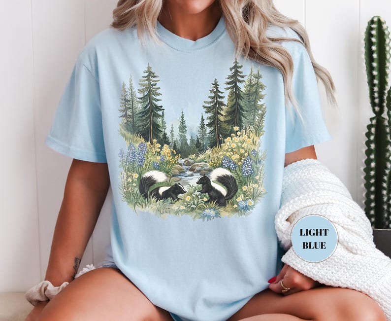 May include: Light blue t-shirt featuring a nature scene with two skunks near a stream, surrounded by wildflowers and trees. The shirt has a relaxed fit and a crew neck. The text "LIGHT BLUE" is on the shirt.