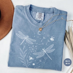 May include: Blue jean colored t-shirt with a white dragonfly and bee graphic. The shirt has a crew neck and short sleeves. A gold necklace with a pendant is draped across the shirt. The shirt is made of a soft, comfortable material.