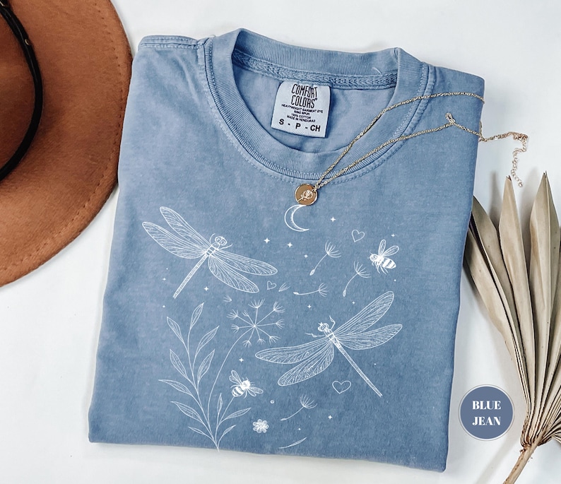 May include: Blue jean colored t-shirt with a white dragonfly and bee graphic. The shirt has a crew neck and short sleeves. A gold necklace with a pendant is draped across the shirt. The shirt is made of a soft, comfortable material.