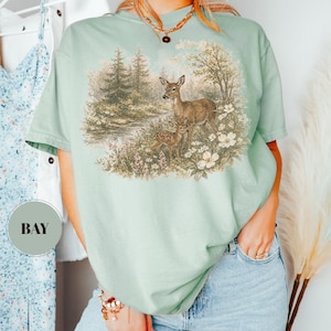May include: A light green t-shirt featuring a nature scene with deer and a fawn in a forest setting. The graphic includes trees, flowers, and a small body of water. The shirt is a comfortable, casual style.