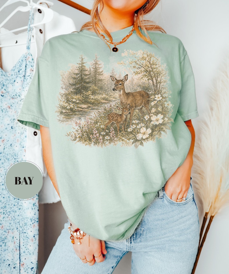 May include: A light green t-shirt featuring a nature scene with deer and a fawn in a forest setting. The graphic includes trees, flowers, and a small body of water. The shirt is a comfortable, casual style.