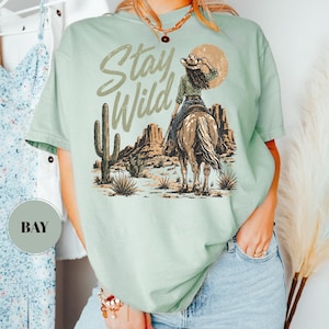 May include: A pale green t-shirt featuring the text "Stay Wild" above a desert landscape illustration. The design includes a person on a horse, a cactus, and a sun. The t-shirt has a loose fit.