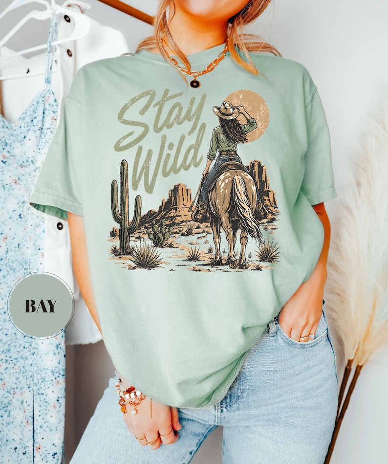 May include: A pale green t-shirt featuring the text "Stay Wild" above a desert landscape illustration. The design includes a person on a horse, a cactus, and a sun. The t-shirt has a loose fit.