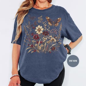 May include: A denim blue short-sleeve t-shirt featuring a graphic print of a wildflower bouquet in shades of cream, brown, and red, accented by two butterflies. A small circular badge on the lower right side contains the text DENIM.