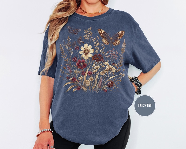 May include: A denim blue short-sleeve t-shirt featuring a graphic print of a wildflower bouquet in shades of cream, brown, and red, accented by two butterflies. A small circular badge on the lower right side contains the text DENIM.