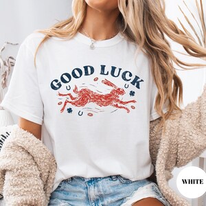 K&ouml;nnte beinhalten: Wei&szlig;es T-Shirt mit dem Text "GOOD LUCK" in Marineblau, bogenf&ouml;rmig angeordnet. Ein roter Hase rennt darunter, mit marineblauen Hufeisen und Kleebl&auml;ttern. Ein l&auml;ssiges Kleidungsst&uuml;ck f&uuml;r den Alltag.