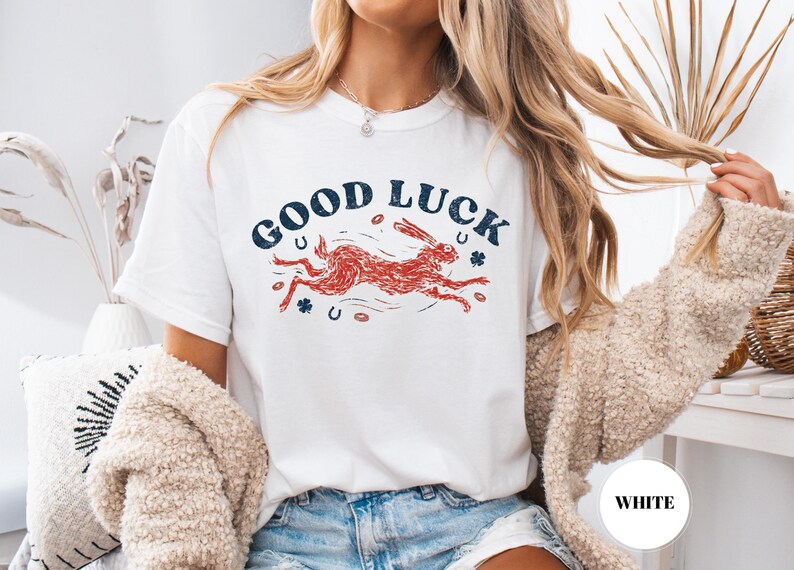 K&ouml;nnte beinhalten: Wei&szlig;es T-Shirt mit dem Text "GOOD LUCK" in Marineblau, bogenf&ouml;rmig angeordnet. Ein roter Hase rennt darunter, mit marineblauen Hufeisen und Kleebl&auml;ttern. Ein l&auml;ssiges Kleidungsst&uuml;ck f&uuml;r den Alltag.