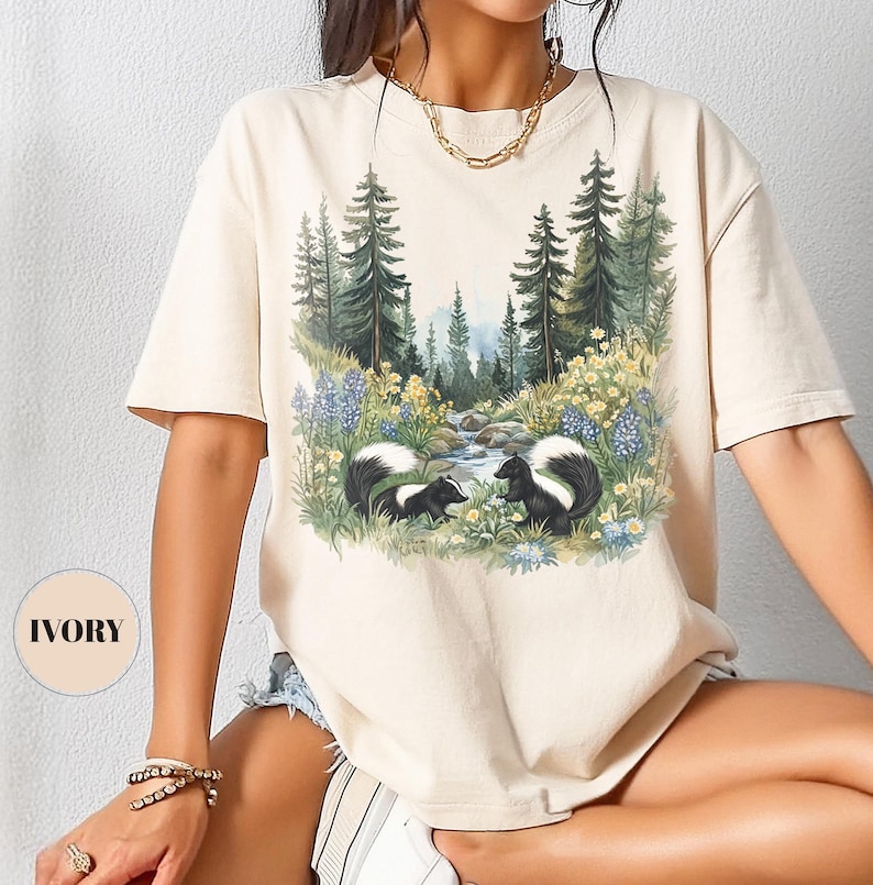 May include: Ivory-colored t-shirt featuring a nature scene with two skunks, a stream, and trees. The graphic includes blue and yellow wildflowers. The shirt has short sleeves and a relaxed fit. The word "IVORY" is printed on a round label.