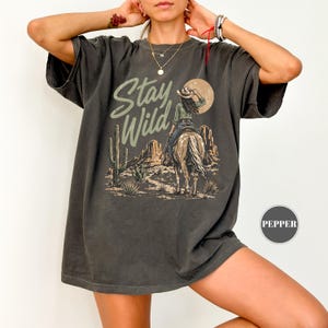 May include: Dark grey t-shirt with a desert landscape design. It features a cowgirl on horseback, cacti, and the words "Stay Wild". The design includes a sun and mountain range.