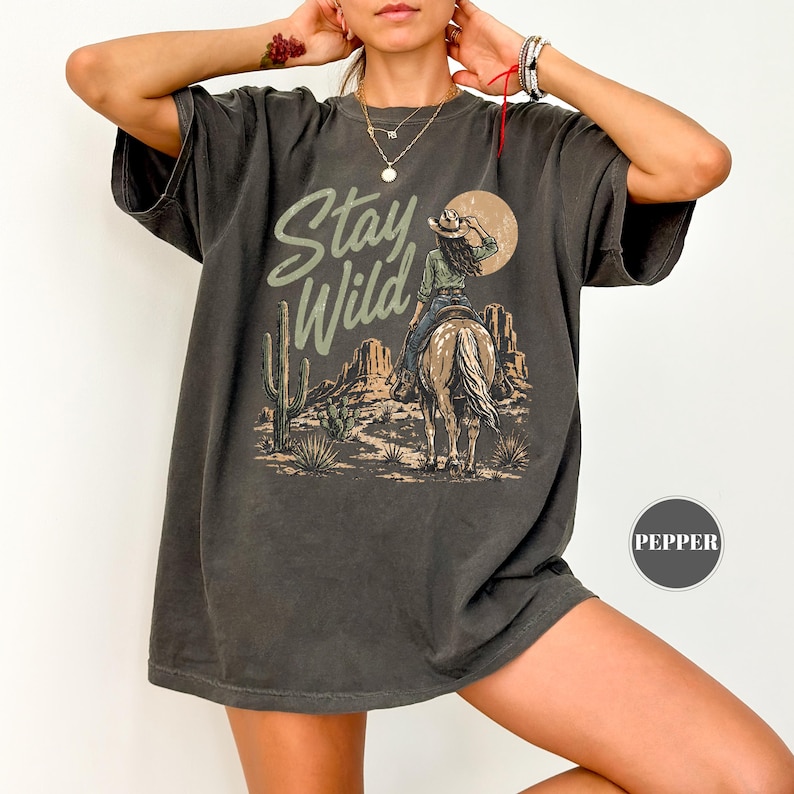 May include: Dark grey t-shirt with a desert landscape design. It features a cowgirl on horseback, cacti, and the words "Stay Wild". The design includes a sun and mountain range.