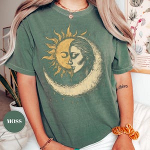 May include: A moss green t-shirt featuring a sun and moon graphic. The sun is depicted with rays and a closed eye, while the moon shows a woman's face. The design is in shades of yellow and cream.