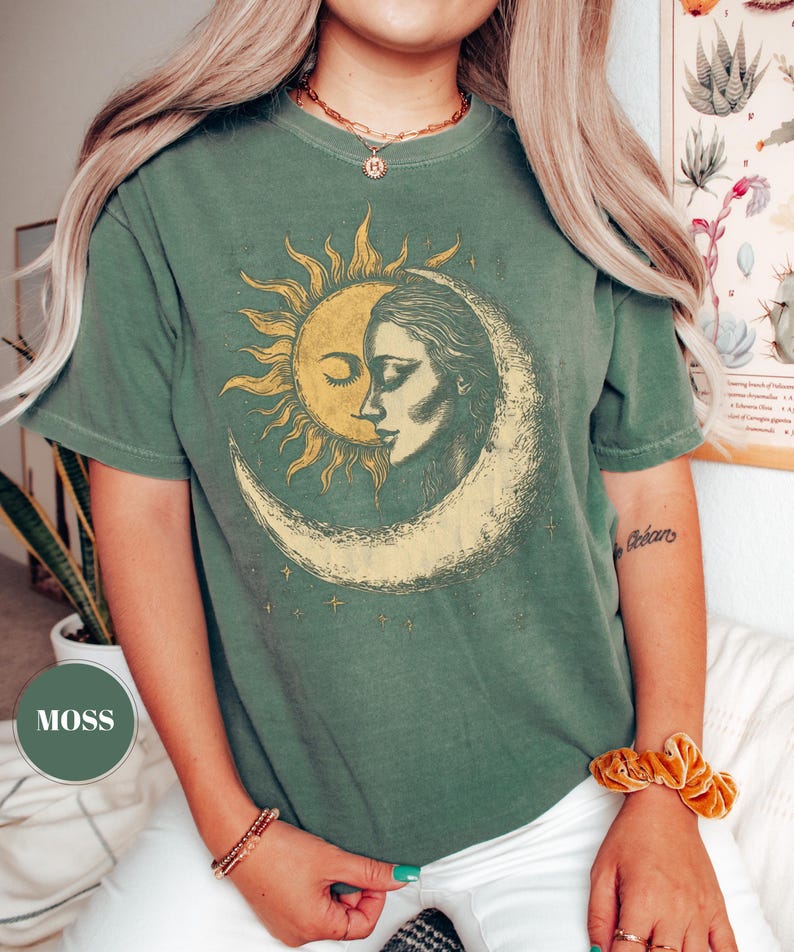 May include: A moss green t-shirt featuring a sun and moon graphic. The sun is depicted with rays and a closed eye, while the moon shows a woman's face. The design is in shades of yellow and cream.
