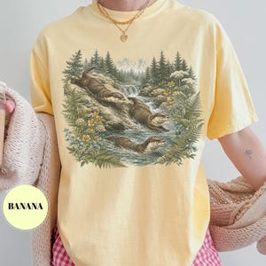 Puede incluir: Una camiseta de manga corta de color amarillo p&aacute;lido con un estampado gr&aacute;fico detallado de tres nutrias nadando en un arroyo forestal. La ilustraci&oacute;n incluye pinos, helechos, flores silvestres y una peque&ntilde;a cascada. La palabra BANANA est&aacute; impresa en negro.