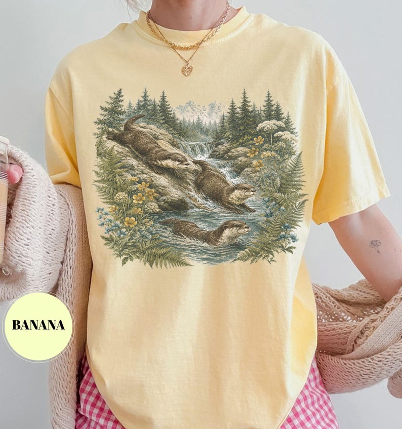 Puede incluir: Una camiseta de manga corta de color amarillo p&aacute;lido con un estampado gr&aacute;fico detallado de tres nutrias nadando en un arroyo forestal. La ilustraci&oacute;n incluye pinos, helechos, flores silvestres y una peque&ntilde;a cascada. La palabra BANANA est&aacute; impresa en negro.