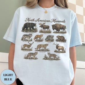 May include: A light blue t-shirt featuring a graphic print titled North American Mammals. The design displays illustrations of an American black bear, moose, bison, gray wolf, red fox, raccoon, beaver, white-tailed deer, mountain lion, elk, coyote, and bobcat.