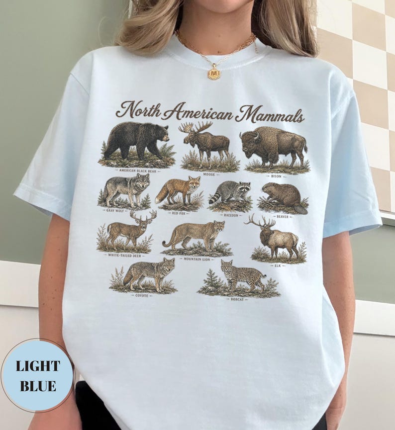 May include: A light blue t-shirt featuring a graphic print titled North American Mammals. The design displays illustrations of an American black bear, moose, bison, gray wolf, red fox, raccoon, beaver, white-tailed deer, mountain lion, elk, coyote, and bobcat.