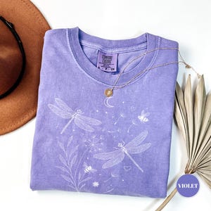 May include: A violet-colored t-shirt with a crew neck, featuring a white dragonfly and floral design. The shirt has a Comfort Colors tag and is paired with a gold necklace. A brown hat is visible in the upper left corner.