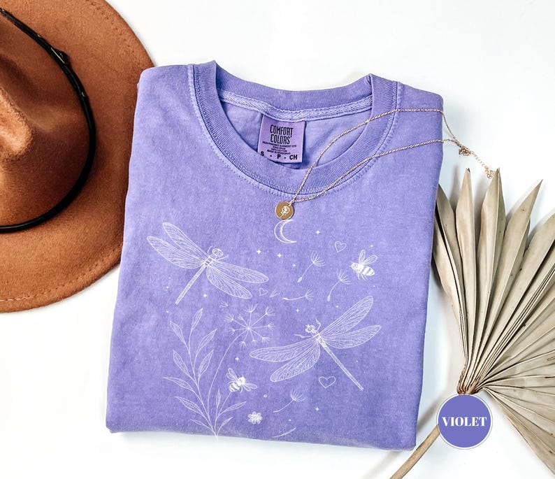May include: A violet-colored t-shirt with a crew neck, featuring a white dragonfly and floral design. The shirt has a Comfort Colors tag and is paired with a gold necklace. A brown hat is visible in the upper left corner.