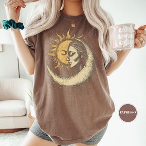 May include: Brown t-shirt featuring a celestial design with a sun and moon graphic. The graphic has a vintage aesthetic. The shirt is worn with gray shorts. A mug with the text "Cool Mom Club" is held in the hand.