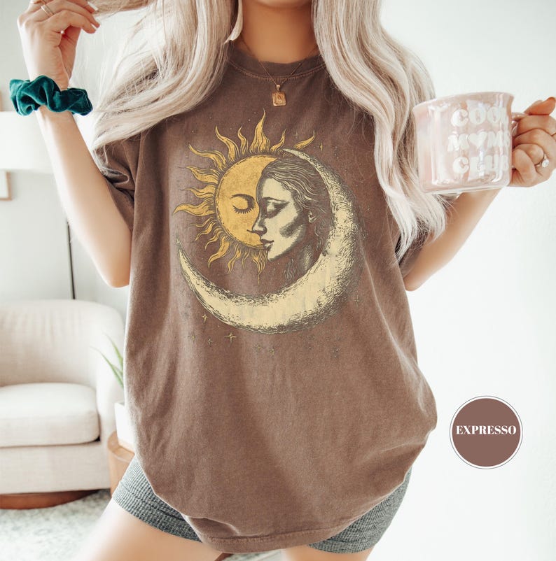 May include: Brown t-shirt featuring a celestial design with a sun and moon graphic. The graphic has a vintage aesthetic. The shirt is worn with gray shorts. A mug with the text "Cool Mom Club" is held in the hand.