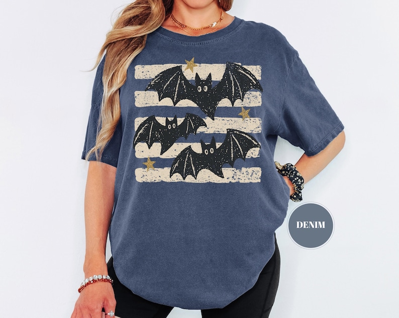 May include: A denim blue short-sleeve t-shirt featuring a graphic print of three black bats flying across horizontal distressed beige stripes. Small gold-colored stars are interspersed between the bats. A circular label reads DENIM.