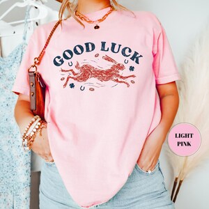K&ouml;nnte beinhalten: Hellrosa T-Shirt mit dem Schriftzug "GOOD LUCK" in Marineblau, einer laufenden Hasenillustration, Hufeisen und Kleebl&auml;ttern. Das T-Shirt hat eine hellrosa Farbe. Ein l&auml;ssiges Kleidungsst&uuml;ck.