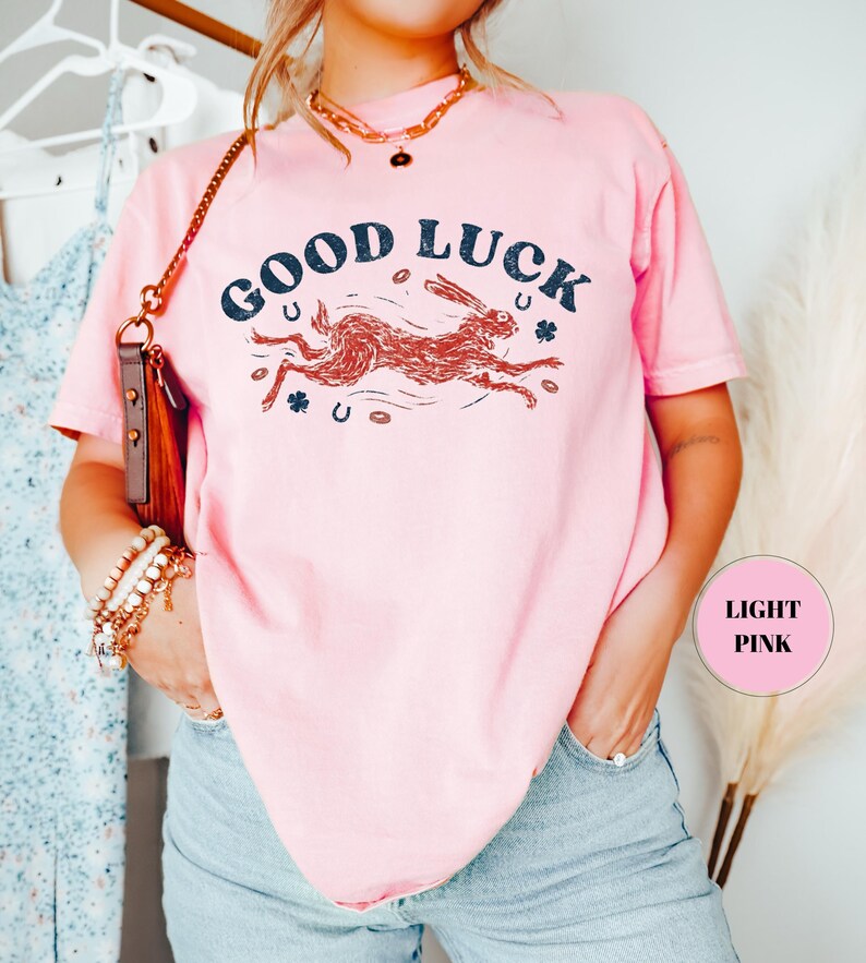 K&ouml;nnte beinhalten: Hellrosa T-Shirt mit dem Schriftzug "GOOD LUCK" in Marineblau, einer laufenden Hasenillustration, Hufeisen und Kleebl&auml;ttern. Das T-Shirt hat eine hellrosa Farbe. Ein l&auml;ssiges Kleidungsst&uuml;ck.